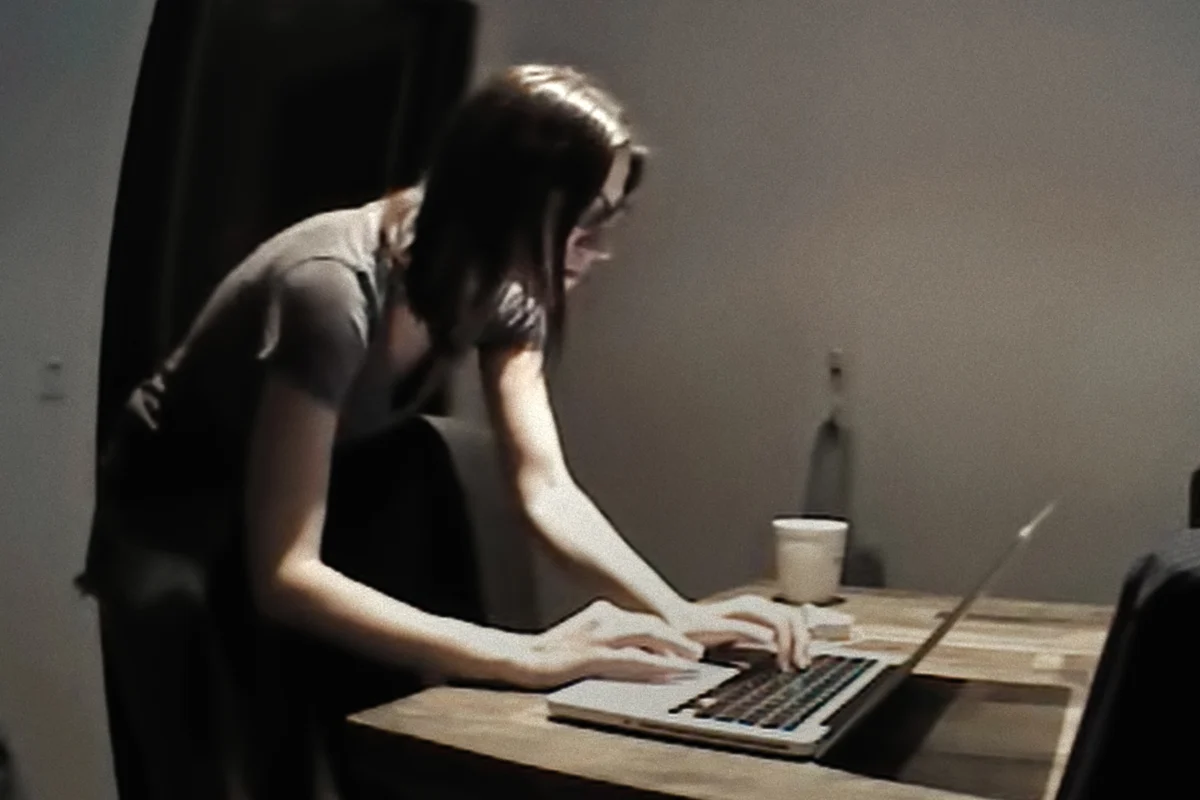 "Woman standing and typing on a laptop at a wooden table in a dimly lit room with a cup and chairs nearby."